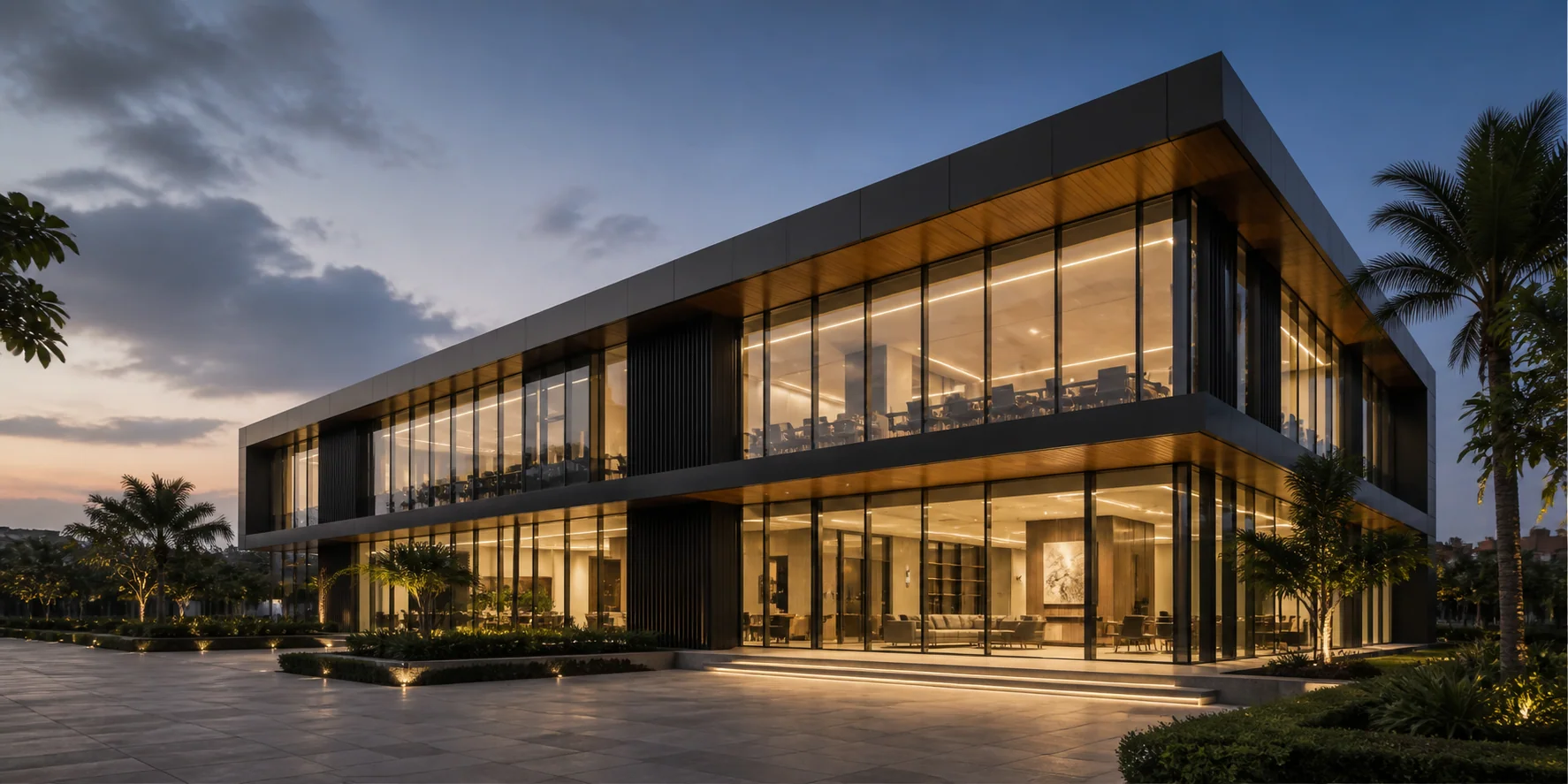A contemporary two-storey glass and timber building lit at dusk, with landscaped plaza and palms